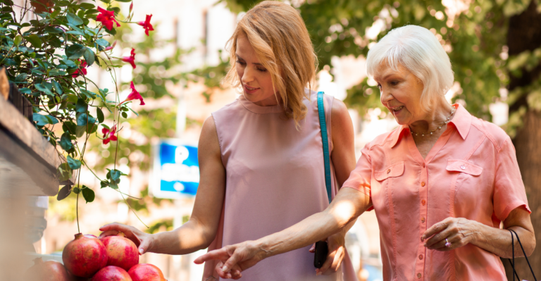 A younger woman and an older woman smile while selecting fresh pomegranates at an outdoor market, symbolizing healthy eating and caregiving support.