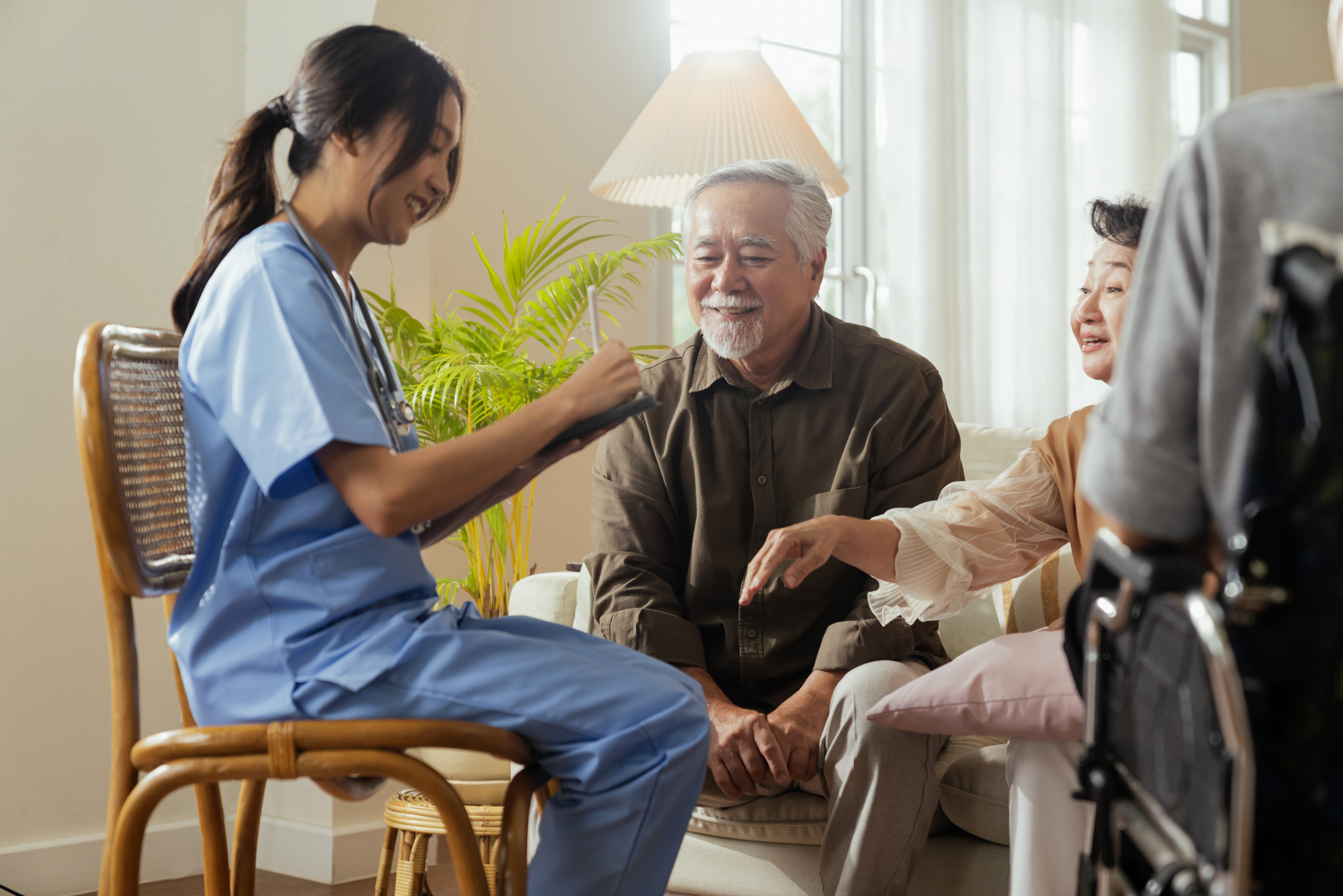 Smiling in-home caregiver in blue scrubs takes notes while speaking with an elderly couple in their living room, with a wheelchair nearby, highlighting compassionate elderly care and personalized in-home care services.