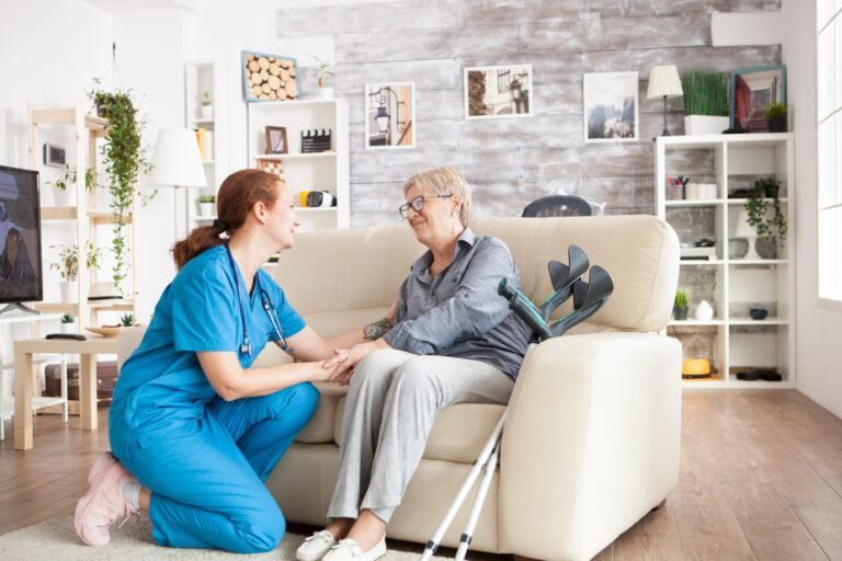 A caregiver in blue scrubs kneels beside an elderly woman seated on a cream sofa, holding her hand and smiling warmly in a cozy, plant-filled living room.